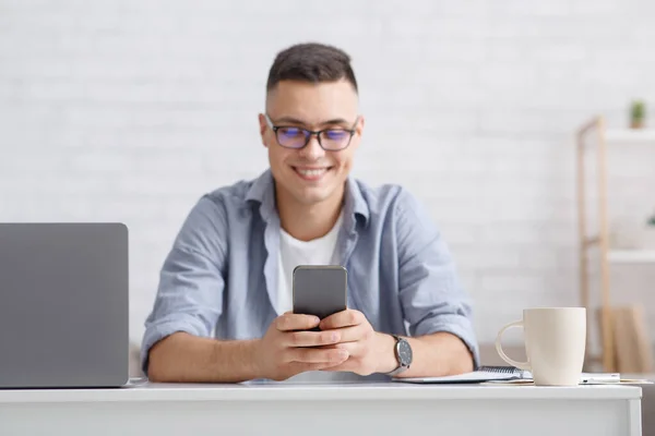 Chatear con seguidores o grandes comentarios y comentarios. Sonriente joven en gafas de escribir en el teléfono inteligente en el lugar de trabajo con el ordenador portátil en el interior de la sala de estar — Foto de Stock