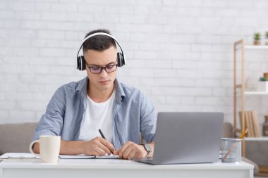Covid-19 salgını sırasında evde öğrenci ve eğitim. Gözlüklü ve modern kulaklıklı meşgul bir adam deftere not alır ve dizüstü bilgisayarla online ders izler.