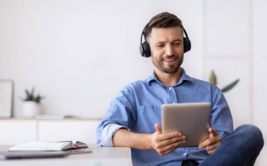Kulaklık takan yakışıklı iş adamı iş yerinde dijital tabletle dinleniyor ve dinleniyor.
