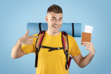 Sırt çantalı, uluslararası pasaport ve uçak biletlerini mavi stüdyo arka planında gösteren olumlu milenyum adamı.