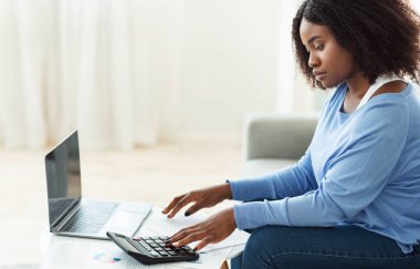 Evde hesap makinesi ve dizüstü bilgisayar kullanan siyah bir kadın.
