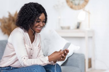 Mutlu siyah kadın evde akıllı telefondan müzik dinliyor, kulaklık takıyor.