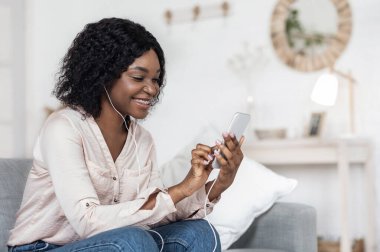 Akıllı telefondan interneti karıştıran ve evde müzik dinleyen mutlu Afrikalı kadın.