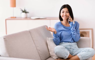 Gülümseyen Asyalı kadın koltukta oturmuş cep telefonuyla konuşuyor.