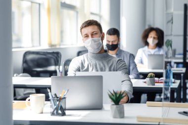 Koruyucu maske takan milenyum erkek çalışanı salgın sırasında iş yerinde koruyucu camla doküman ve dizüstü bilgisayarla çalışıyor.