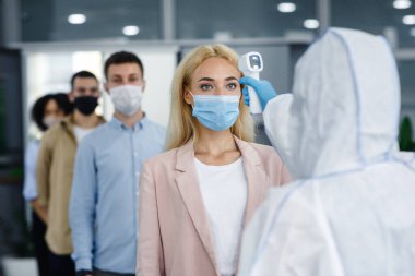 Coronavirüs salgını sırasında kurumsal sosyal mesafeler, sağlığı korumak için yeni bir standarttır. Kızıl ötesi termometresi olan tehlikeli madde giysisi giyen adam insanların sıcaklığını kontrol ediyor.