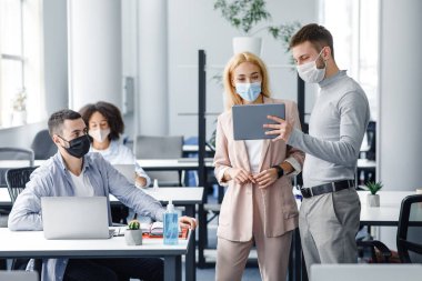 Şirket şirketinde takım çalışması ve koronavirüs salgını sırasında sosyal mesafe. Koruyucu maskeli erkek yönetici, içerideki işçilere tablet gösteriyor