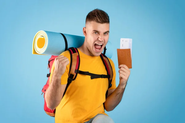 Guapo turista masculino sosteniendo pasaporte con boletos de avión y gritando de emoción en fondo azul estudio — Foto de Stock