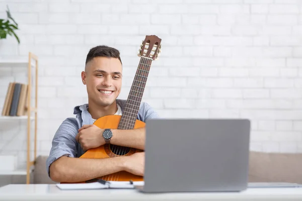 Nuovo hobby durante l'autoisolamento, app per l'educazione e la chitarra. Uomo felice abbraccia la chitarra e guarda laptop a casa interna durante l'epidemia di coronavirus — Foto Stock