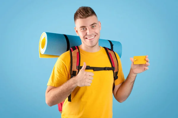 Presupuesto de viaje. Cool millennial chico con mochila y tarjeta de crédito que muestra el pulgar hacia arriba gesto sobre fondo de estudio azul — Foto de Stock