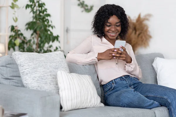 Hushållsvila. Glad ung svart kvinna Avkopplande på soffan med smartphone — Stockfoto