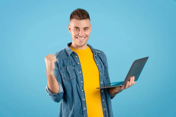 Online ταξιδιωτικό γραφείο. Θετικός νεαρός με laptop κράτηση εισιτηρίων στο διαδίκτυο και κάνοντας ΝΑΙ χειρονομία πάνω από το μπλε φόντο — Φωτογραφία Αρχείου