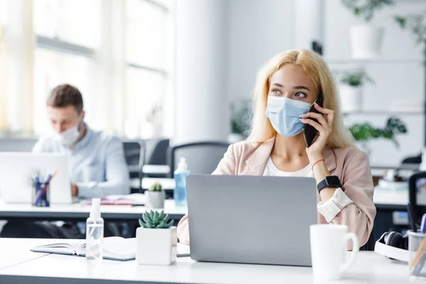 Terug aan het werk na covid-19 quarantaine. Bezig met millennial vrouw in beschermende masker praten over de telefoon en typen op laptop op het werk met antiseptische in moderne kantoor interieur — Stockfoto