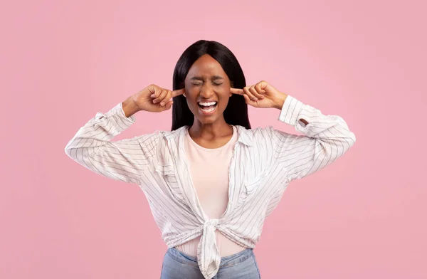 Retrato da jovem negra irritada fechando as orelhas com os dedos e fechando os olhos no fundo do estúdio rosa — Fotografia de Stock