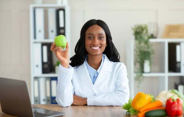 Dietista femenina negra positiva sosteniendo fruta de manzana y mirando la cámara en la clínica —  Fotos de Stock