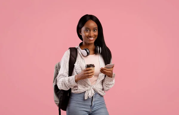 Mujer bastante negra con teléfono inteligente y auriculares que sostienen el café para llevar en el fondo del estudio rosa — Foto de Stock