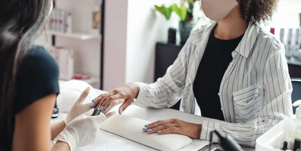 Perfecte nagels en manicure proces. Professionele manicure in handschoenen bedekt blauwe nagels met top tot Afrikaans amerikaanse klant in beschermende masker — Stockfoto