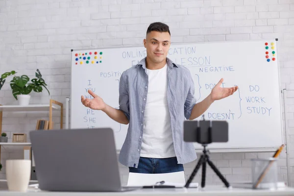 Online class and English lesson at home during covid-19. Millennial man spreads his arms, communicates with students and pupils on laptop and smartphone in interior of living room