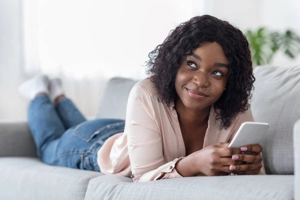 Mulher africana Pensivo usando smartphone enquanto deitado no sofá confortável em casa — Fotografia de Stock
