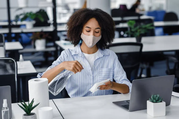 Higiene no local de trabalho para proteger a saúde contra o coronavírus. Africano americano mulher em máscara usar spray e papel toalhas para desinfectar mesa e laptop no interior do escritório — Fotografia de Stock
