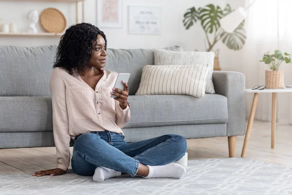 Millennial Black Donna seduta con smartphone sul pavimento a casa, guardando lontano — Foto Stock