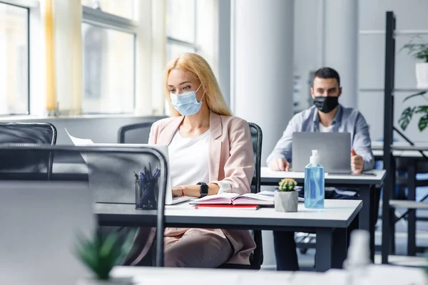 New working conditions and social distancing after covid-19 quarantine. Blonde woman in protective mask works with documents and laptop