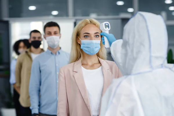 Corporate social distance are new standard to protect health during coronavirus epidemic. Man in hazmat suit with infrared thermometer checks temperature of people — Stock Photo, Image