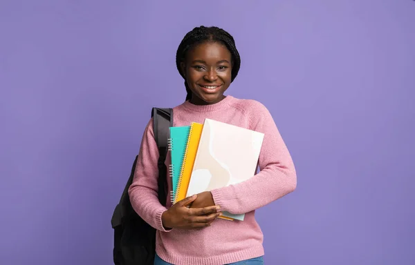 Estudiante negra sonriente con libros de trabajo y mochila posando sobre fondo morado — Foto de Stock