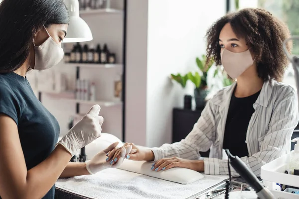 Nieuw normaal en strijd tegen het virus in schoonheidssalon. aziatische vrouwelijke meester in beschermende masker werkt met nagels en desinfecteren handen met antiseptische van afrikaanse amerikaanse dame — Stockfoto