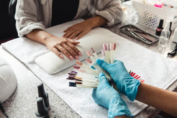 Wybierz modny kolor w studiu kosmetycznym. Afroamerykanka w rękawiczkach ochronnych pokazujących próbki lakieru do paznokci do klienta na stole ze sprzętem do paznokci podczas covid-19 — Zdjęcie stockowe