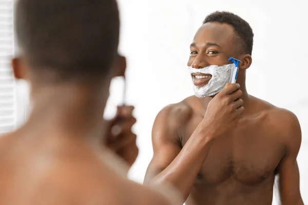 Alegre negro chico afeitado cara de pie cerca espejo en cuarto de baño — Foto de Stock