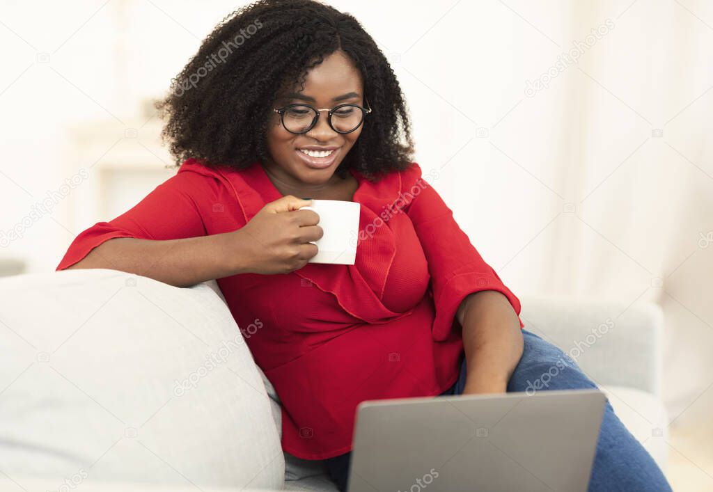 Black woman using laptop drinking coffee sitting on sofa