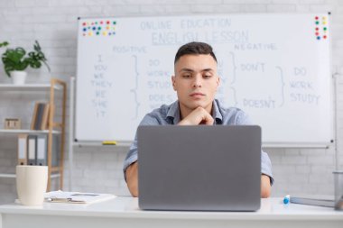 COVID-19 salgını sırasında öğretmeninizle İngilizce çalışın. Odaklanmış genç erkek öğretmen internetten öğrenciyi dinliyor ve oturma odasındaki dizüstü bilgisayara bakıyor.