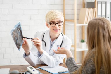 Klinikteki doktora danışıyorum. Beyaz önlüklü ve gözlüklü yetişkin kadın terapist röntgen filmi gösteriyor.