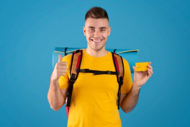 Seyahat bütçesi. Turist vitesli çekici genç adam başparmağını kaldırıyor ve mavi arka planda kredi kartını gösteriyor.