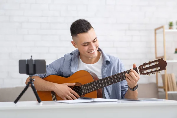 Молодой студент учится играть на гитаре во время самоизоляции дома или онлайн-репетитора. Улыбающийся парень смотрит на гитару и делает видео уроки для последователей в гостиной во время вспышки вируса — стоковое фото