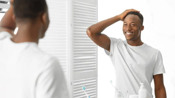 Hombre africano guapo tocando el pelo de pie cerca del espejo en el baño — Foto de Stock