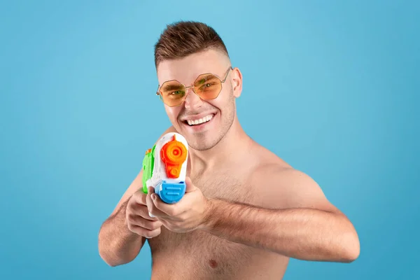 Conceito de férias de verão. Atraente homem milenar apontando arma de água na tela sobre o fundo do estúdio azul — Fotografia de Stock