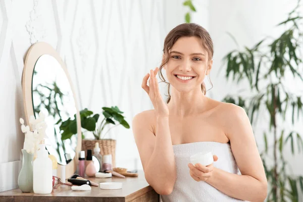 Atractiva joven mujer usando crema facial en el dormitorio —  Fotos de Stock