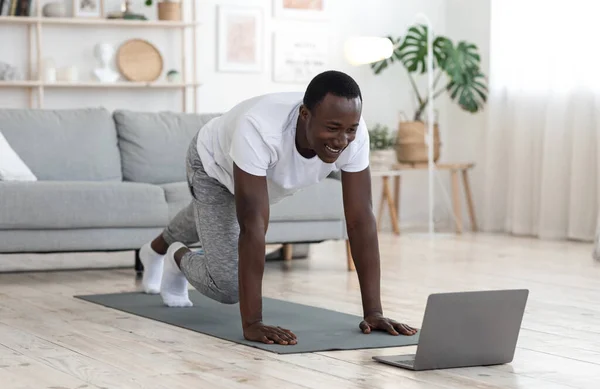 Alegre chico negro haciendo ejercicio en casa, mirando la pantalla del ordenador portátil — Foto de Stock