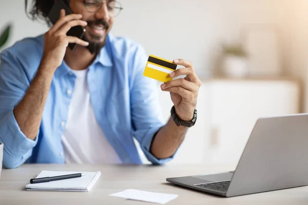 Pago móvil. Hombre indio positivo sosteniendo tarjeta de crédito y hablando por teléfono celular — Foto de Stock