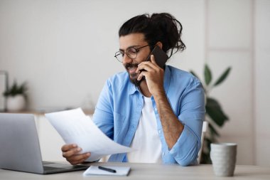 Hintli serbest çalışan adam cep telefonuyla konuşuyor ve ofiste evraklarla çalışıyor.