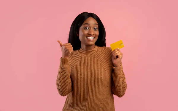 Mulher negra feliz segurando cartão de crédito e mostrando polegar para cima gesto no fundo do estúdio rosa — Fotografia de Stock