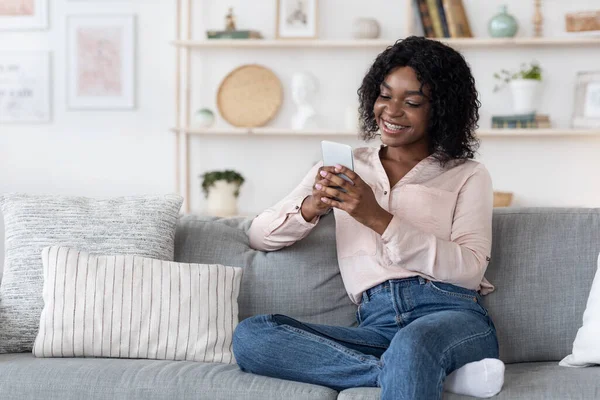 Wesoły czarny kobieta relaks na kanapie w salonie z telefonem komórkowym — Zdjęcie stockowe