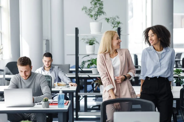 Volviendo al trabajo y comunicándome después del cierre. Felices mujeres europeas y afroamericanas chismorrean en el interior de la oficina moderna — Foto de Stock