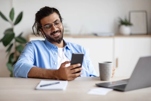 Empleado indio sonriente relajarse con teléfono inteligente y café en la oficina — Foto de Stock