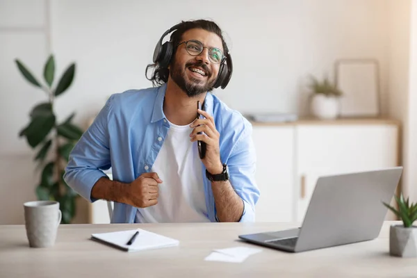 Alegre indio freelancer chico cantando y escuchando música con auriculares mientras trabaja —  Fotos de Stock