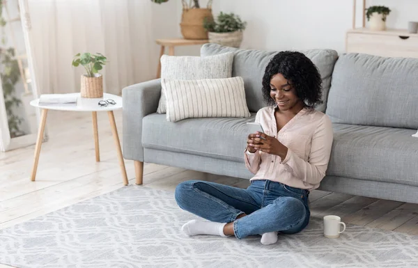 Wesoły czarny kobieta relaks w domu ze smartfonem i kawy — Zdjęcie stockowe