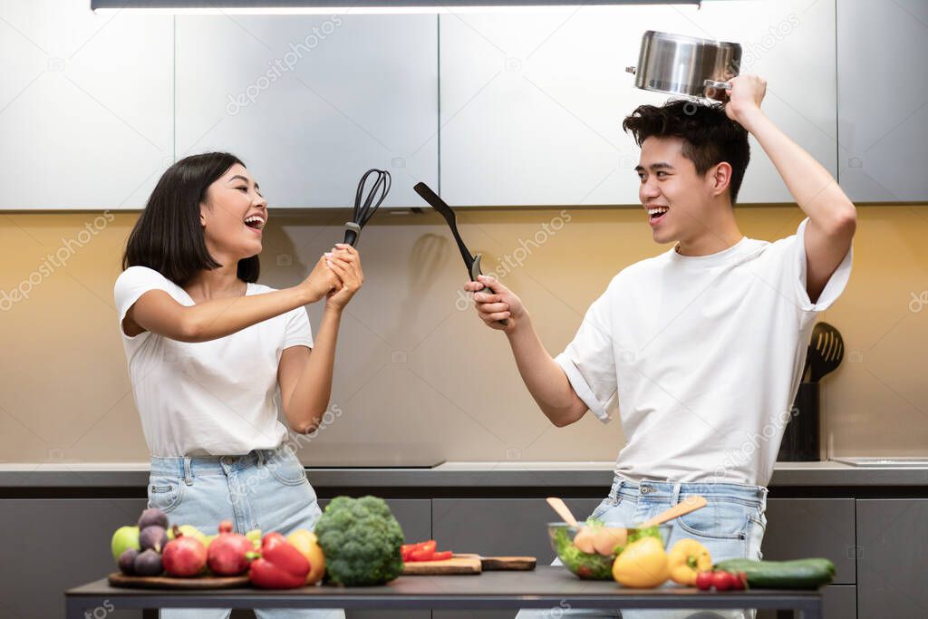 Asian Couple Fencing Holding Cooking Tools Having Fun In Kiitchen