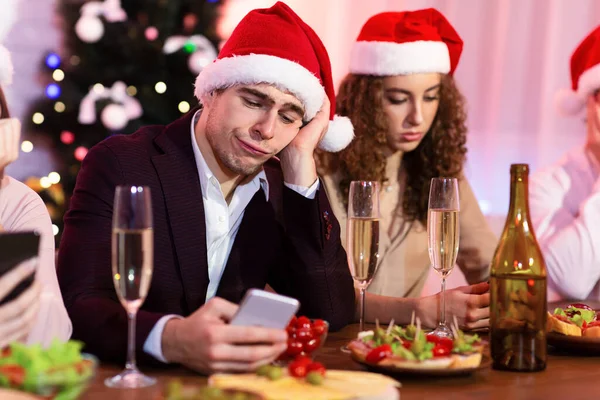 Unatkozó fickó használ okostelefon alatt karácsonyi party barátaival beltéri — Stock Fotó